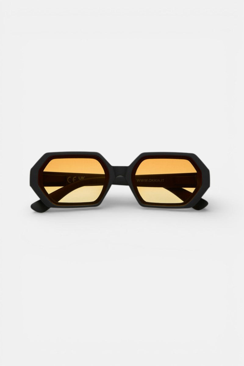 Eine schwarze hexagonale Sonnenbrille von Okkia mit bernsteinfarbenen Gläsern wird auf einem weißen Hintergrund gezeigt. Der dicke, matte Rahmen und der UV-400-Schutz sorgen für eine stilvolle Abdeckung, wobei sowohl die Gläser als auch der Steg deutlich sichtbar sind.