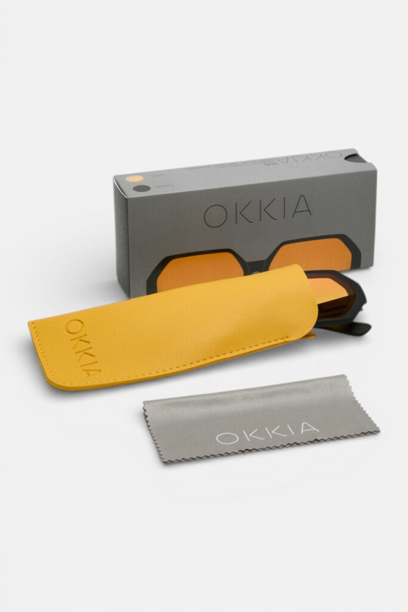 Eine Okkia-Sonnenbrille mit orangefarbenen Gläsern und geometrischem Design sitzt teilweise in einem gelben Etui vor einer grauen Okkia-Box. Ein graues OKKIA-Reinigungstuch liegt daneben, alles auf weißem Hintergrund.