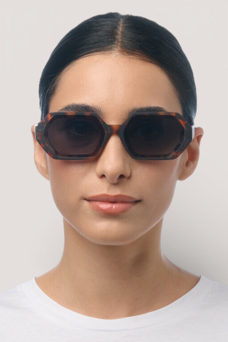 Eine Frau mit dunklem, nach hinten gezogenem Haar trägt eine große, sechseckige Okkia Andrea Sonnenbrille mit UV 400 Schutz und ein schlichtes weißes Hemd und blickt mit neutralem Gesichtsausdruck nach vorne auf einen hellgrauen Hintergrund.