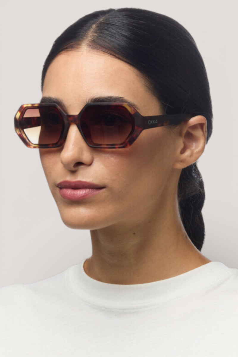 Eine Frau mit langem, dunklem Haar trägt die übergroße sechseckige Sonnenbrille von Okkia mit UV-400-Schutz und ein weißes Oberteil mit Rundhalsausschnitt. Sie posiert neutral vor einem schlichten hellgrauen Hintergrund und blickt leicht nach links.