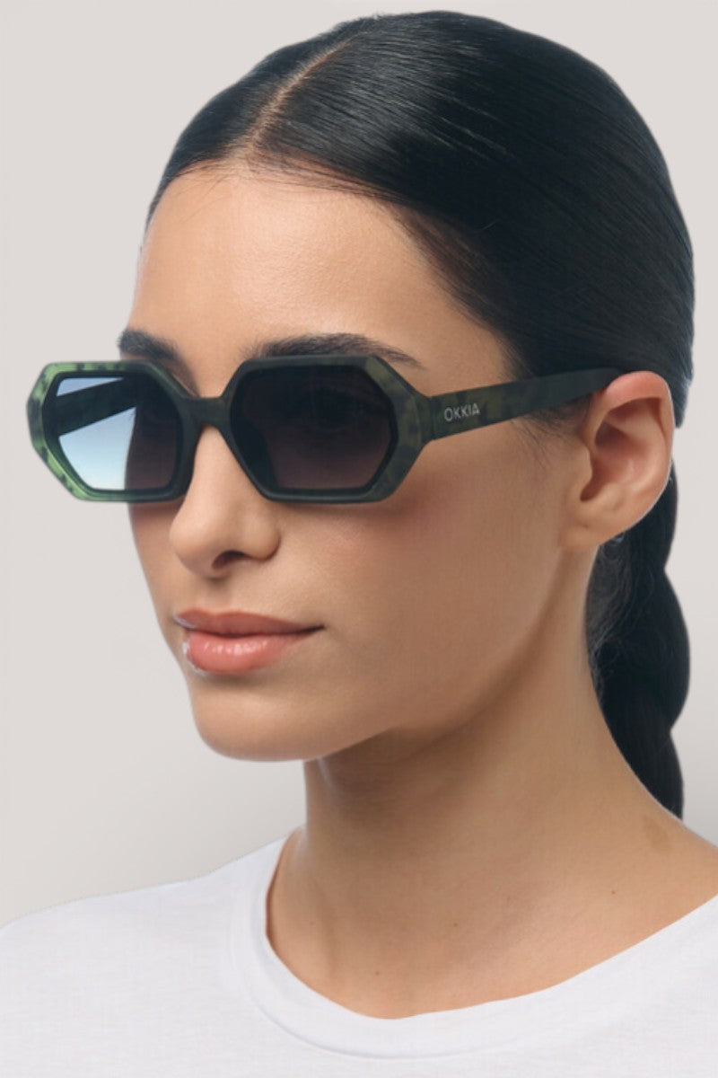 Eine Frau mit langem, dunklem Haar trägt die geometrische sechseckige Sonnenbrille von Okkia mit schwarzen Bügeln und UV-400-Schutz. Sie hat glatte, helle Haut, ein neutrales Make-up und ein schlichtes weißes Oberteil vor einem hellgrauen Hintergrund.