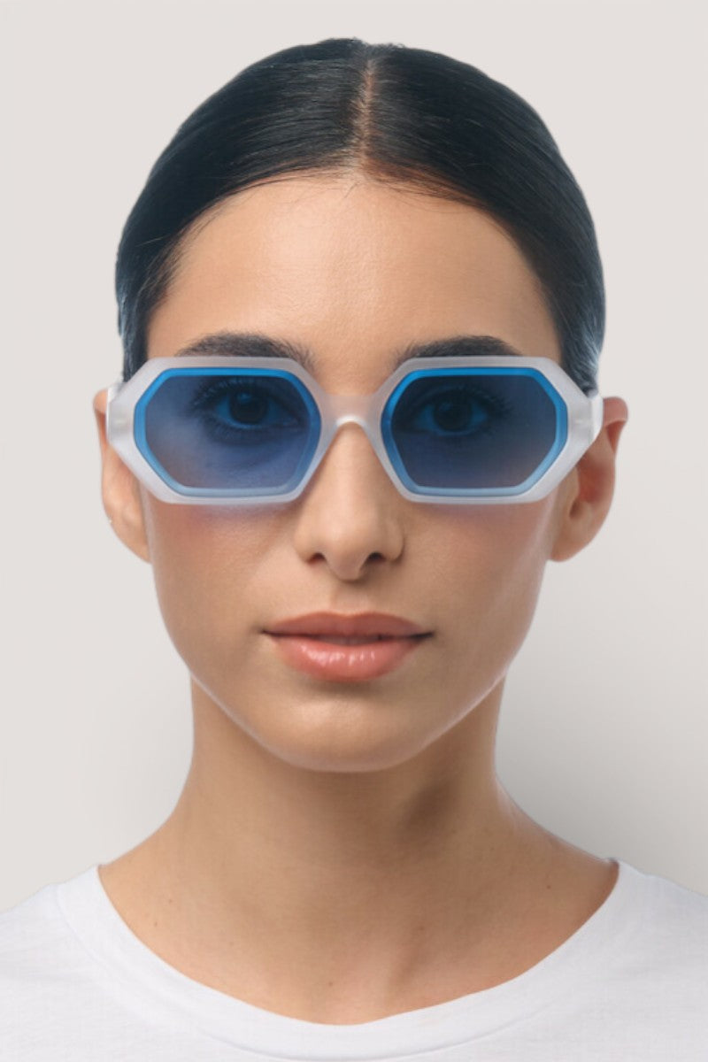 Eine Frau mit glattem, dunklem Haar, das in der Mitte gescheitelt ist, trägt eine große blaue sechseckige Sonnenbrille mit weißem Rahmen von Okkia. Sie schaut in einem schlichten weißen Oberteil vor einem hellen Hintergrund nach vorne, ihr Gesichtsausdruck ist neutral.