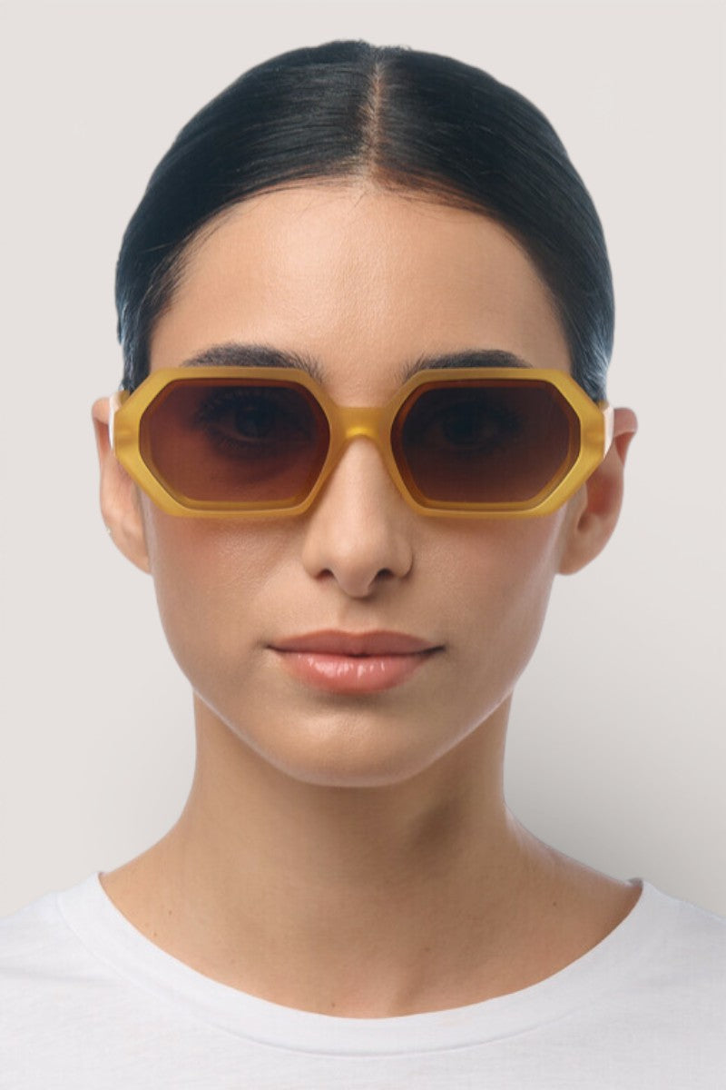 Eine Person mit dunklem, nach hinten gezogenem Haar, die eine gelbe geometrische Sonnenbrille von Okkia Andrea mit braunen Gläsern und UV-400-Schutz trägt, blickt vor einem schlichten hellen Hintergrund nach vorne. Ihr Gesichtsausdruck ist neutral.