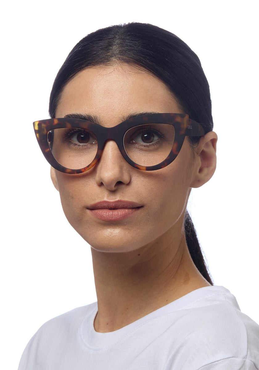 A woman with straight, dark hair tied back wears large, cat-eye Schildpattmuster Lesebrille from Okkia and a white shirt. She faces forward with a neutral expression against a plain white background.