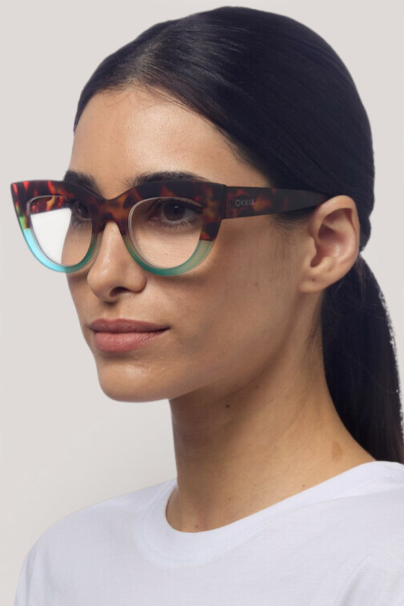 Eine Frau mit langen dunklen Haaren, die nach hinten gebunden sind, trägt eine auffällige Okkia Damenbrille im Cat-Eye-Design mit einem Schildpatt-Muster auf der Oberseite und durchscheinenden tealfarbenen Rändern darunter. Sie hat eine klare Haut, gepflegte Augenbrauen und trägt ein schlichtes weißes Hemd vor einem neutralen Hintergrund.