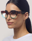 Eine Frau mit langen dunklen Haaren, die nach hinten gebunden sind, trägt eine auffällige Okkia Damenbrille im Cat-Eye-Design mit einem Schildpatt-Muster auf der Oberseite und durchscheinenden tealfarbenen Rändern darunter. Sie hat eine klare Haut, gepflegte Augenbrauen und trägt ein schlichtes weißes Hemd vor einem neutralen Hintergrund.