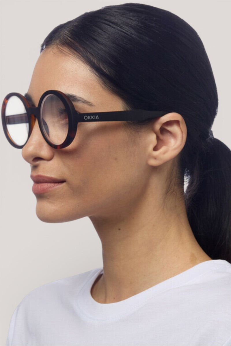 Eine Frau mit glattem schwarzem Haar in einem niedrigen Pferdeschwanz trägt eine große, runde Okkia-Lesebrille mit einem übergroßen Design und dicken Schildpattrahmen. Sie ist mit einem schlichten weißen Oberteil bekleidet und von der Seite vor einem hellgrauen Hintergrund dargestellt.