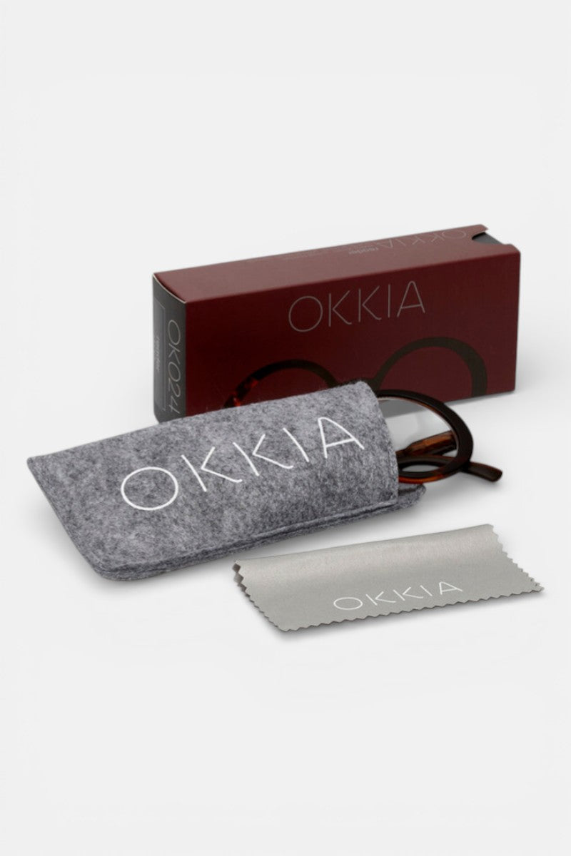 Eine Okkia-Brille in Übergröße, teilweise in einem grauen Okkia-Filzbeutel, sitzt vor einer kastanienbraunen Okkia-Schachtel, daneben ein passendes graues Okkia-Putztuch - alles auf einem weißen Hintergrund.