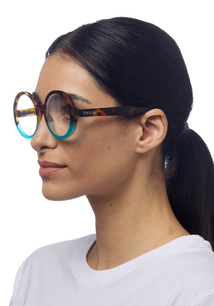 A woman with straight dark hair in a low ponytail wears large, round Okkia Lesebrille in an oversize design that fade to blue at the edges. She is shown in profile facing left, wearing a white T-shirt against a plain white background.