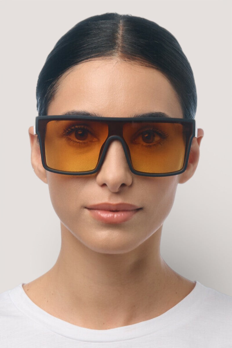 Eine Person mit dunklem, geglättetem Haar, die ein weißes Hemd und eine übergroße Okkia rechteckige Sonnenbrille mit orangefarbenen Gläsern und schwarzem Rahmen trägt, blickt vor einem schlichten hellen Hintergrund direkt in die Kamera.