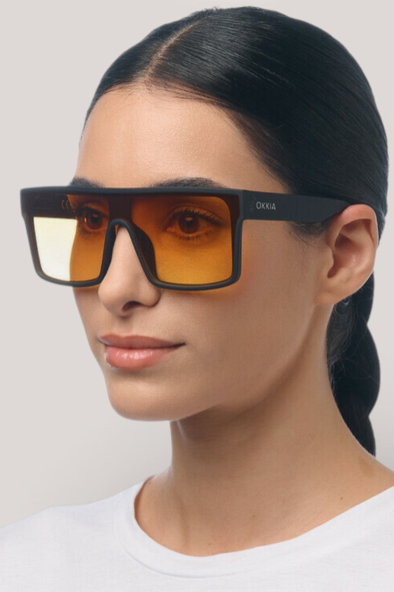 Eine Frau mit glattem, zurückgebundenem dunklem Haar trägt die Oversized rechteckige Sonnenbrille von Okkia mit schwarzem Rahmen und bernsteinfarbenen Gläsern (UV 400 Schutz). Sie hat eine glatte Haut, ein neutrales Make-up, ein schlichtes weißes Hemd und einen hellgrauen Hintergrund.