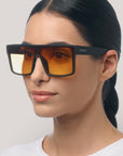 Eine Frau mit glattem, zurückgebundenem dunklem Haar trägt die Oversized rechteckige Sonnenbrille von Okkia mit schwarzem Rahmen und bernsteinfarbenen Gläsern (UV 400 Schutz). Sie hat eine glatte Haut, ein neutrales Make-up, ein schlichtes weißes Hemd und einen hellgrauen Hintergrund.