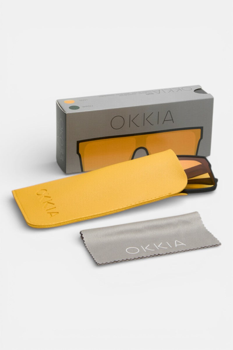 Eine Schachtel mit der Aufschrift "Okkia" und dem Bild einer gelben Brille steht neben der Okkia UV 400 Schutz rechteckige Sonnenbrille, einem gelben Etui und einem grauen Reinigungstuch, die alle ordentlich auf einem weißen Hintergrund angeordnet sind.
