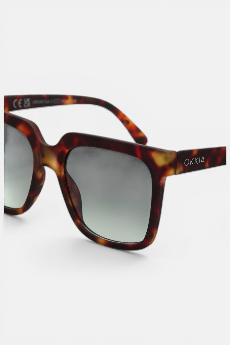 Nahaufnahme einer übergroßen, quadratischen Okkia-Sonnenbrille mit Schildpattmuster, getönten Gläsern mit Farbverlauf, UV-400-Schutz und dem Schriftzug OKKIA" in Weiß auf dem linken Bügel. Auf einfarbig weißem Hintergrund abgebildet, um jedes Detail hervorzuheben.