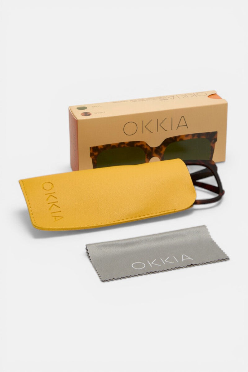 Eine übergroße, quadratische Okkia Sonnenbrille mit UV 400 Schutz befindet sich teilweise in einem gelben Etui, neben einem grauen Reinigungstuch und einer beigen Okkia-Box auf einer weißen Fläche.