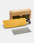 Eine übergroße, quadratische Okkia Sonnenbrille mit UV 400 Schutz befindet sich teilweise in einem gelben Etui, neben einem grauen Reinigungstuch und einer beigen Okkia-Box auf einer weißen Fläche.