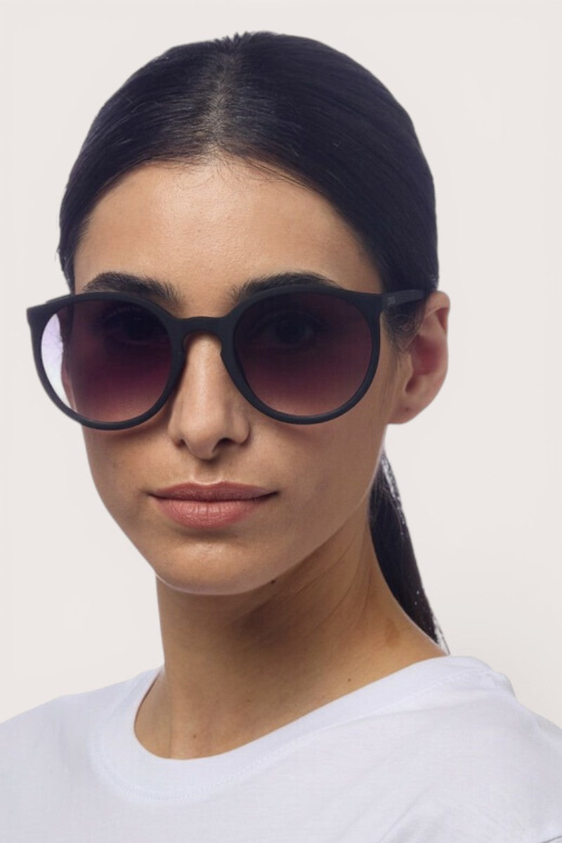 Eine Frau mit glattem, dunklem Haar, das sie sich zusammengebunden hat, trägt eine schwarze Okkia-Sonnenbrille im Retro-Design mit UV-400-Schutz und ein weißes T-Shirt. Vor einem hellen Hintergrund blickt sie leicht nach links.