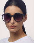 Eine Frau mit glattem, dunklem Haar, das sie sich zusammengebunden hat, trägt eine schwarze Okkia-Sonnenbrille im Retro-Design mit UV-400-Schutz und ein weißes T-Shirt. Vor einem hellen Hintergrund blickt sie leicht nach links.