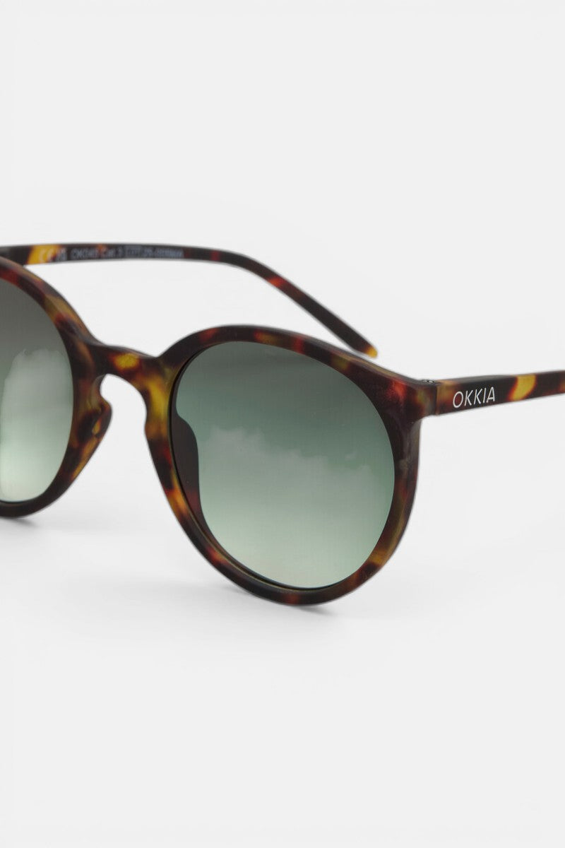Nahaufnahme der Okkia Berlino Sonnenbrille mit abgerundeten Gläsern, grüner Verlaufstönung und braunem Schildpattgestell. Der Schriftzug "OKKIA" erscheint in Weiß auf dem linken Bügel. Mit UV-400-Schutz. Schlichter weißer Hintergrund.