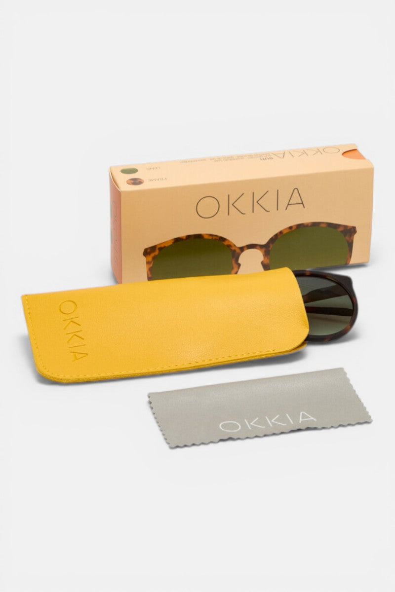 Eine Retro-Sonnenbrille von Okkia mit schildpattfarbenem Rahmen liegt teilweise in einem gelben Etui. Daneben stehen eine pfirsichfarbene Okkia-Schachtel mit Sonnenbrillenmotiv und ein graues Okkia-Putztuch, alles auf weißem Hintergrund.