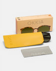 Eine Retro-Sonnenbrille von Okkia mit schildpattfarbenem Rahmen liegt teilweise in einem gelben Etui. Daneben stehen eine pfirsichfarbene Okkia-Schachtel mit Sonnenbrillenmotiv und ein graues Okkia-Putztuch, alles auf weißem Hintergrund.
