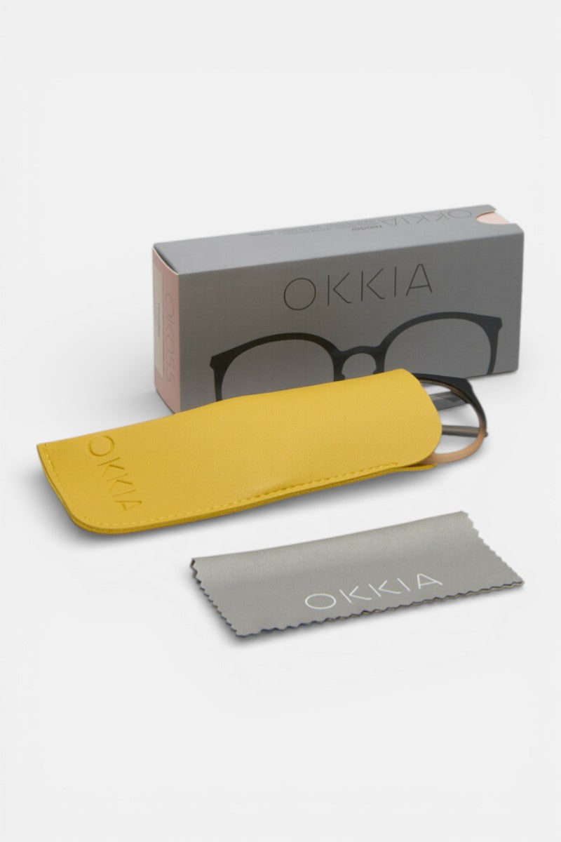 Eine graue Okkia-Brillenbox, ein gelbes Etui mit Soft-Touch-Oberfläche und geprägtem Okkia-Logo, eine teilweise sichtbare Brille im Inneren und ein gefaltetes graues Putztuch mit Okkia-Branding - angeordnet auf weißem Hintergrund.
