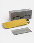 Eine graue Okkia-Brillenbox, ein gelbes Etui mit Soft-Touch-Oberfläche und geprägtem Okkia-Logo, eine teilweise sichtbare Brille im Inneren und ein gefaltetes graues Putztuch mit Okkia-Branding - angeordnet auf weißem Hintergrund.