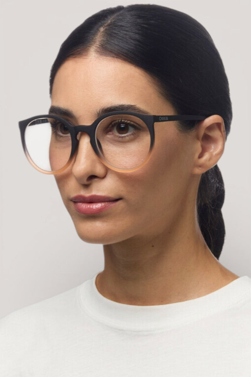 Eine Frau mit glattem, zurückgebundenem dunklem Haar trägt große, runde, schwarz-beige Lesebrille von Okkia und ein weißes Oberteil. Sie blickt leicht nach rechts und hat einen neutralen Ausdruck vor einem schlichten, hellgrauen Hintergrund.