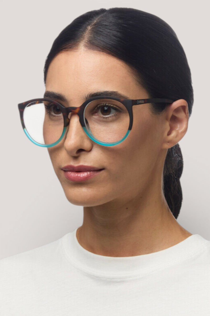 Eine Frau mit langen dunklen Haaren, die nach hinten gebunden sind, trägt eine runde Okkia-Brille in schwarz-grünem Runddesign mit Soft-Touch-Oberfläche, gepaart mit einem weißen Hemd. Sie blickt leicht nach rechts vor einem schlichten hellgrauen Hintergrund, ihr Ausdruck ist neutral.