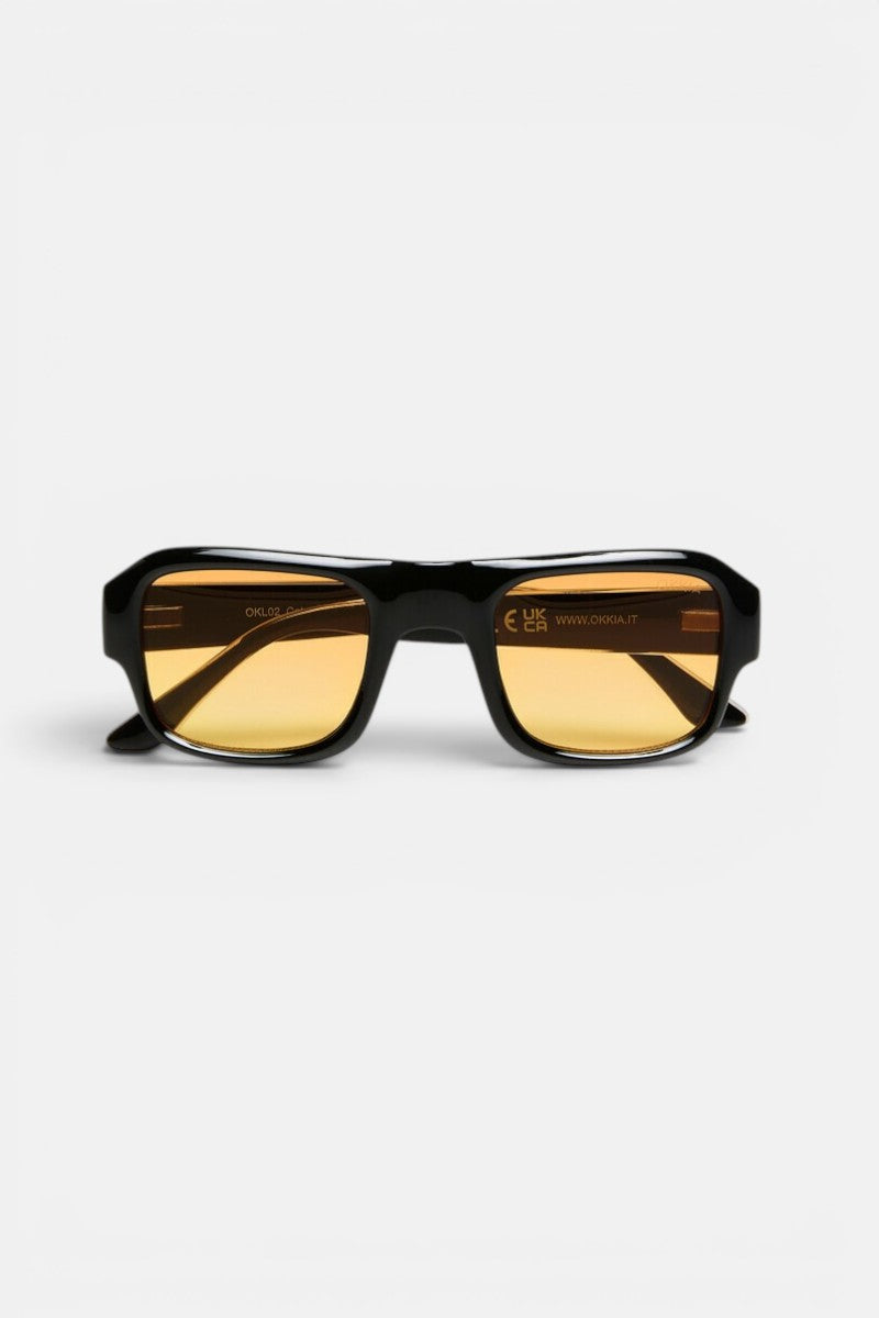 Eine rechteckige Sonnenbrille mit einem breiten, glänzend schwarzen Rahmen und bernsteinfarbenen Gläsern liegt auf einem weißen Hintergrund und ist nach vorne gerichtet. Auf der Innenseite des rechten Glases und des Bügels sind dezente Reflexionen und filigrane Beschriftungen zu erkennen, die den Vintage-Charme dieser Sonnenbrille aus der CLUB-Kollektion von Okkia – Modell STROMBOLI – unterstreichen.