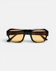 Eine rechteckige Sonnenbrille mit einem breiten, glänzend schwarzen Rahmen und bernsteinfarbenen Gläsern liegt auf einem weißen Hintergrund und ist nach vorne gerichtet. Auf der Innenseite des rechten Glases und des Bügels sind dezente Reflexionen und filigrane Beschriftungen zu erkennen, die den Vintage-Charme dieser Sonnenbrille aus der CLUB-Kollektion von Okkia – Modell STROMBOLI – unterstreichen.