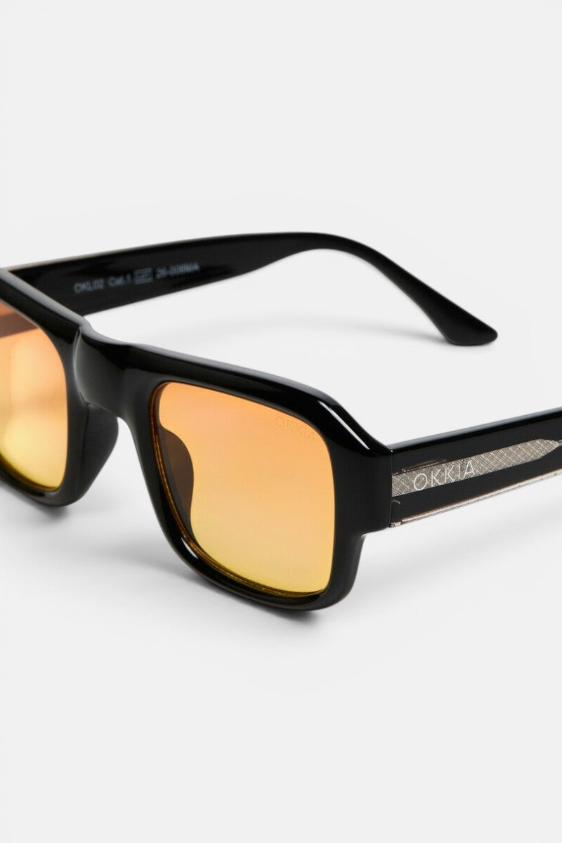 Nahaufnahme einer schwarzen, rechteckigen Sonnenbrille mit orange getönten Spiegelgläsern. Dieses Modell aus der Okkia CLUB-Kollektion im Vintage-Stil verfügt über ein metallisches Detail am Bügel und den Markennamen „Okkia“ in Silber. Der schlichte weiße Hintergrund betont die Reflexion in den Gläsern und unterstreicht das schlanke, moderne Design.