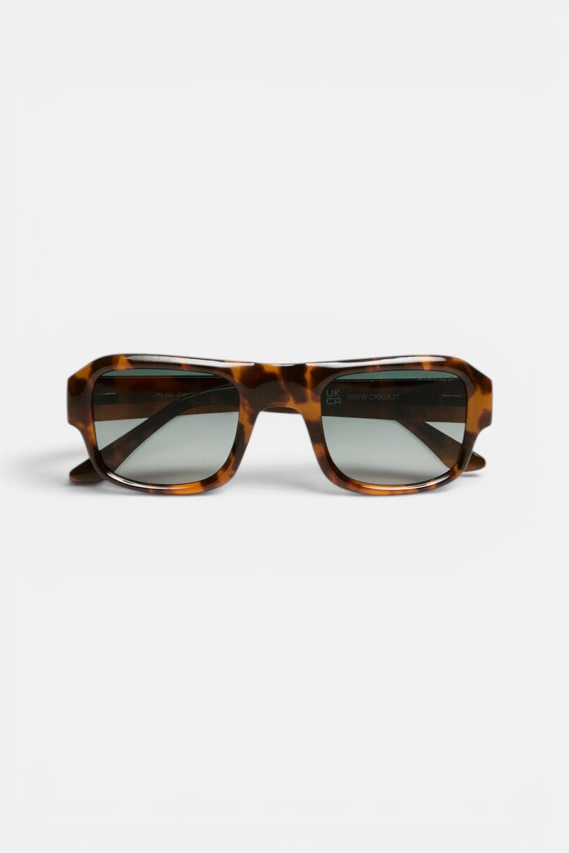 Rechteckige Sonnenbrille im Schildpatt-Look mit dickem, glänzendem Rahmen aus der Okkia STROMBOLI-Kollektion. Die dunklen, leicht verspiegelten Gläser erinnern an den Vintage-Look einer Sonnenbrille. Vor einem schlichten weißen Hintergrund bildet die Sonnenbrille den Blickfang des Bildes, wobei sich die Bügel seitlich nach außen erstrecken.