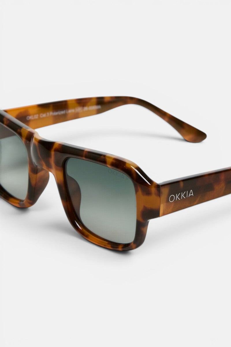 Hier ist Ihr überarbeiteter Satz unter Verwendung der bereitgestellten Produktdaten: Nahaufnahme einer Sonnenbrille aus der Okkia CLUB-Kollektion in Schildpattoptik mit rechteckigen grauen Gläsern. Der glänzende, geometrische Rahmen verleiht ihr einen modernen Touch, während der Markenname „Okkia“ in Weiß auf dem linken Bügel zu sehen ist. Der schlichte weiße Hintergrund unterstreicht das schicke Design dieser STROMBOLI-Sonnenbrille.