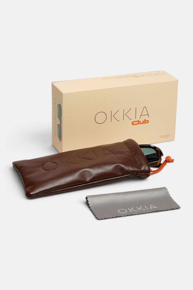 Eine Okkia-Sonnenbrille mit geometrischem Rahmen steckt teilweise in einem braunen, weichen Etui, das neben einer beigen Okkia Club-Schachtel und einem grauen Okkia-Brillenputztuch liegt – alles vor einem weißen Hintergrund angeordnet: ein elegantes Highlight der Vintage-inspirierten Okkia Club-Kollektion.