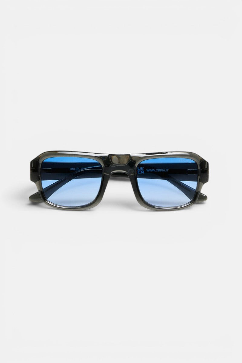 Eine rechteckige Sonnenbrille von Okkia mit durchscheinendem schwarzem Rahmen und blau getönten Gläsern wird vor einem schlichten weißen Hintergrund nach vorne gerichtet präsentiert. Inspiriert von der OKKIA CLUB Kollektion besticht diese stilvolle Sonnenbrille durch einen breiten, geometrischen Rahmen und einen sanft geschwungenen Steg. Das moderne, schlanke Design macht sie zu einem herausragenden Accessoire aus der STROMBOLI-Brillenlinie.