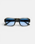 Eine rechteckige Sonnenbrille von Okkia mit durchscheinendem schwarzem Rahmen und blau getönten Gläsern wird vor einem schlichten weißen Hintergrund nach vorne gerichtet präsentiert. Inspiriert von der OKKIA CLUB Kollektion besticht diese stilvolle Sonnenbrille durch einen breiten, geometrischen Rahmen und einen sanft geschwungenen Steg. Das moderne, schlanke Design macht sie zu einem herausragenden Accessoire aus der STROMBOLI-Brillenlinie.