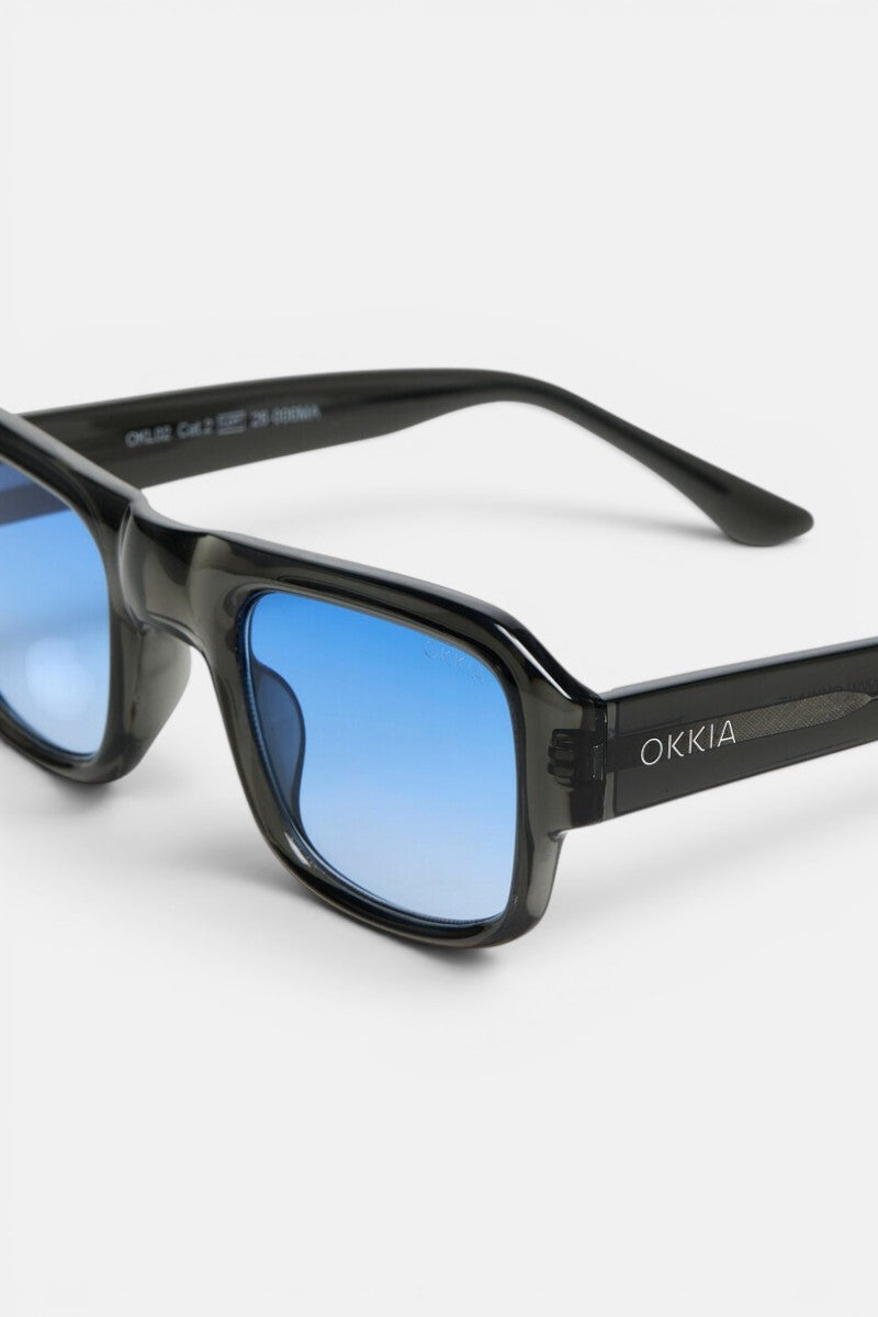 Eine Sonnenbrille mit schwarzem Rahmen und blauen Verlaufsgläsern wird in Nahaufnahme vor einem weißen Hintergrund gezeigt. Auf dem rechten Bügel ist der Markenname „Okkia“ in weißen Buchstaben zu sehen. Diese STROMBOLI-Brille ist Teil der OKKIA CLUB-Kollektion und zeichnet sich durch einen leicht rechteckigen, geometrischen Rahmen sowie eine glänzende Oberfläche aus.