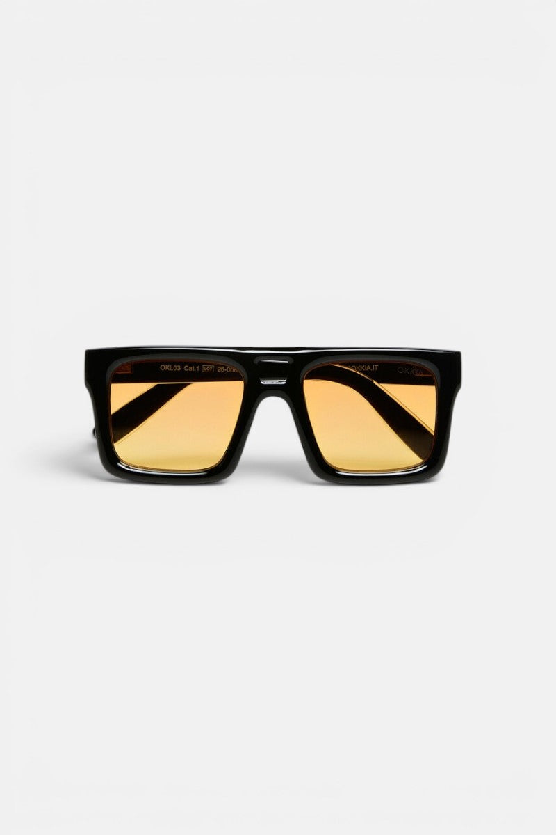 Eine schwarze, rechteckige Sonnenbrille von Okkia mit breitem Rahmen und gelb getönten Gläsern ist auf einem schlichten weißen Hintergrund zu sehen. Die Bügel der Sonnenbrille sind gekreuzt und unterstreichen das markante, moderne Design mit auffälligen Oversize-Proportionen, das für einen wahrhaft zeitgemäßen Look sorgt.