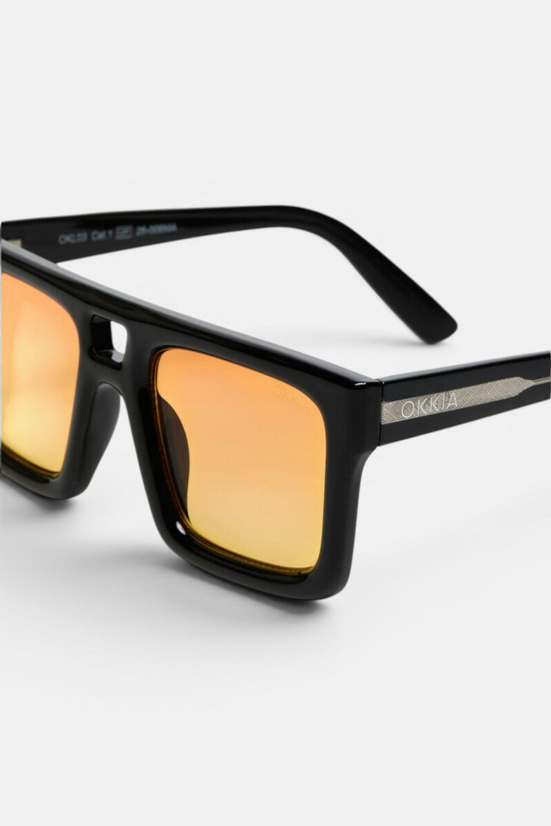 Nahaufnahme der Okkia VULCANO Sonnenbrille: schwarze, rechteckige Sonnenbrille mit goldverspiegelten Gläsern. Der glänzende Rahmen unterstreicht das moderne Design, während der Schriftzug „Okkia“ in Weiß auf den mattierten Bügeln eingraviert ist. Ein hellgrauer Hintergrund hebt das Bild hervor, wobei ein Teil der Innenseite des Bügels und Produktdetails sichtbar sind, was die übergroßen Proportionen dieses stilvollen Accessoires unterstreicht.