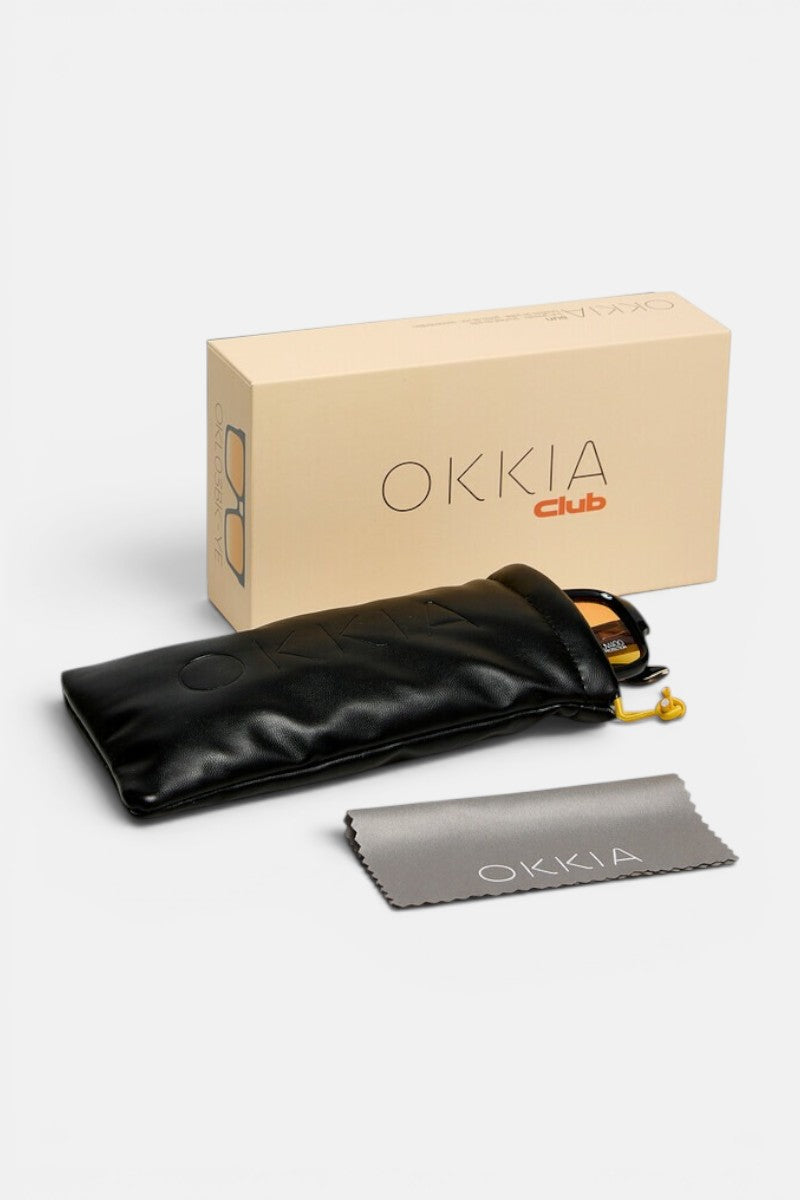Eine beigefarbene Okkia Club-Box ist zusammen mit einer VULCANO-Sonnenbrille zu sehen, die sich durch modernes Design und übergroße Proportionen auszeichnet und teilweise in einem schwarzen Weichetui neben einem grauen Reinigungstuch mit der Aufschrift „Okkia“ zu erkennen ist. Die Artikel sind ordentlich auf einer weißen Fläche angeordnet.