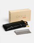 Eine beigefarbene Okkia Club-Box ist zusammen mit einer VULCANO-Sonnenbrille zu sehen, die sich durch modernes Design und übergroße Proportionen auszeichnet und teilweise in einem schwarzen Weichetui neben einem grauen Reinigungstuch mit der Aufschrift „Okkia“ zu erkennen ist. Die Artikel sind ordentlich auf einer weißen Fläche angeordnet.