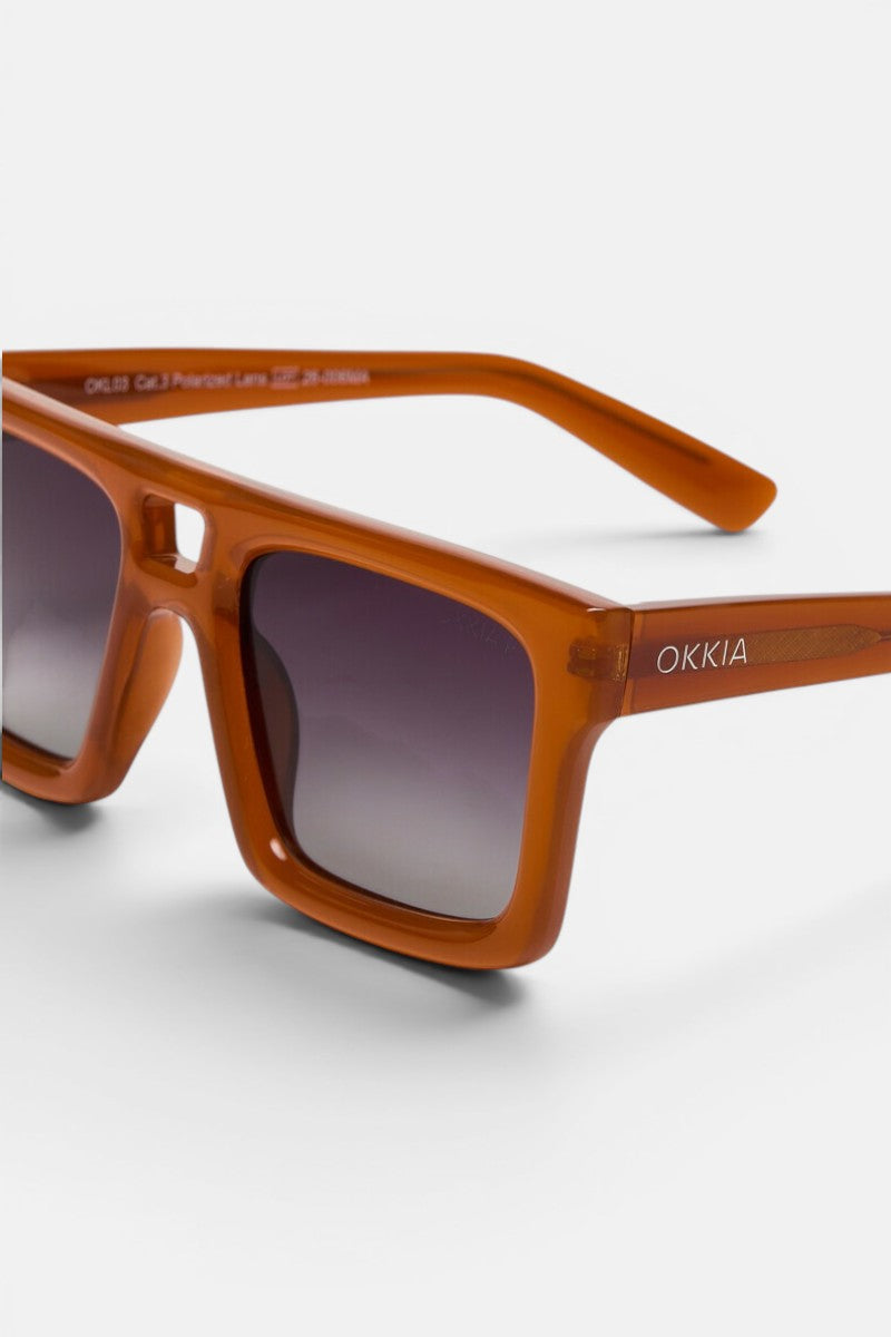 Nahaufnahme einer orangefarbenen, rechteckigen Okkia VULCANO Sonnenbrille mit übergroßem, breitem Rahmen und grauen Verlaufsgläsern. Der Markenname „Okkia“ ist in Weiß auf dem linken Bügel aufgedruckt. Vor einem schlichten, hellen Hintergrund hebt das Bild das moderne Design und die stilvolle Ausstrahlung der Sonnenbrille hervor.