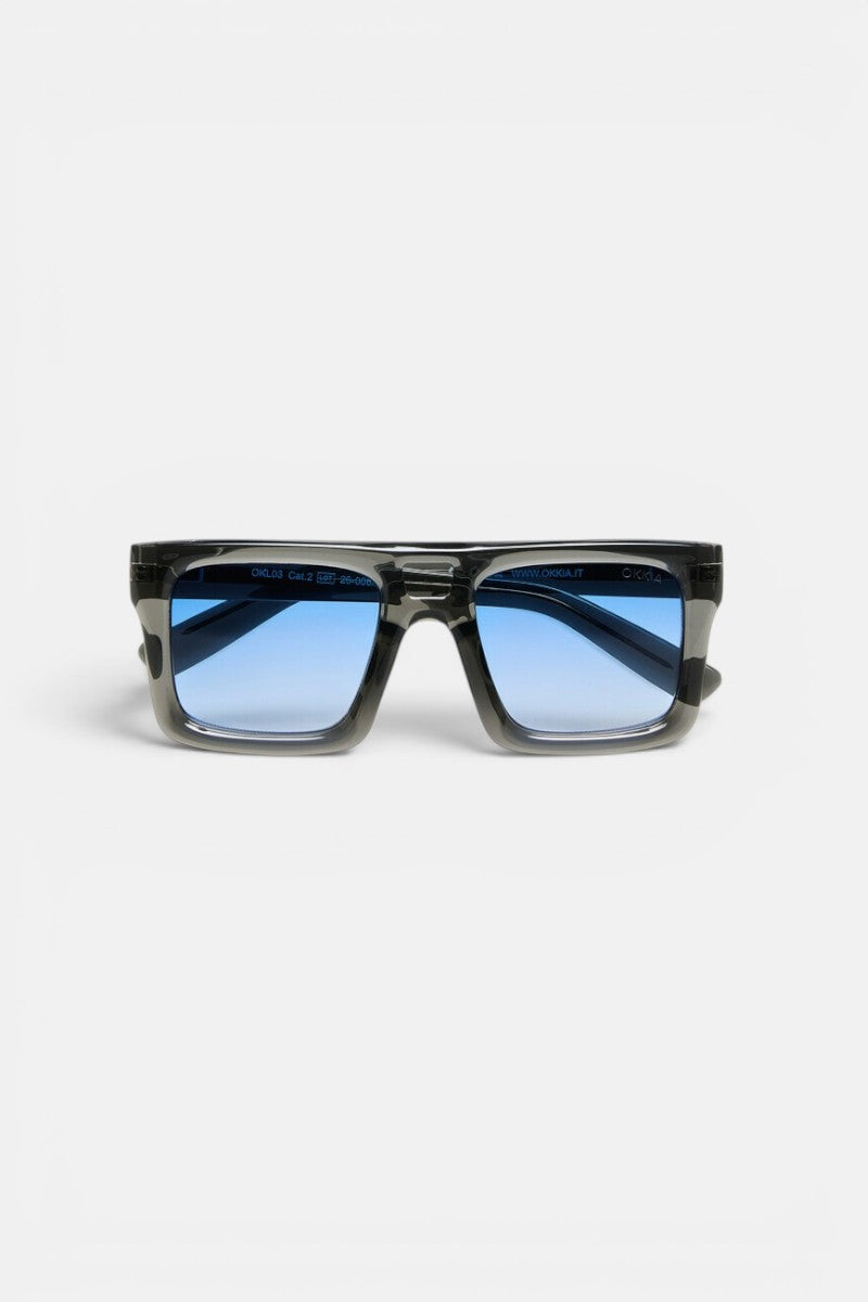 Rechteckige Okkia VULCANO Sonnenbrille mit dickem, schwarz-transparentem Rahmen und blau getönten Gläsern, abgebildet in Frontalansicht vor einem schlichten weißen Hintergrund. Das moderne Design zeichnet sich durch Oversize-Proportionen und Gläser mit Farbverlauf aus, der von tiefem zu hellem Blau übergeht.