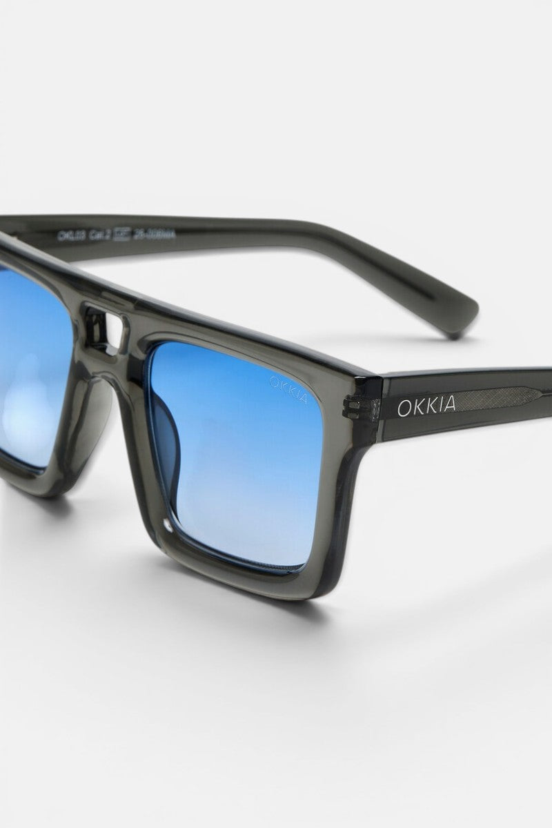 Diese rechteckige Sonnenbrille der Marke Okkia aus der VULCANO-Kollektion besticht durch einen glänzend schwarzen Rahmen und blaue Verlaufsgläser und verkörpert ein modernes Design mit übergroßen Proportionen. Das OKKIA-Logo ist in Weiß auf den linken Bügel aufgedruckt. Die Brille wird vor einem weißen Hintergrund präsentiert, wobei das Bild eng beschnitten ist, um die linke Seite und das Markenlogo hervorzuheben.
