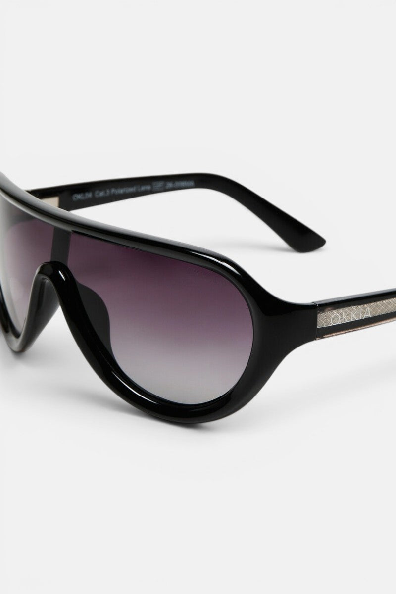 Nahaufnahme einer schwarzen Sonnenbrille von Okkia mit breitem, abgerundetem Rahmen und polarisierten Gläsern in einem Farbverlauf von dunklem zu hellem Violett. Der linke Bügel ist mit einem metallischen Akzent versehen, in den der Markenname eingraviert ist. Vor einem schlichten weißen Hintergrund unterstreicht diese elegante, moderne Sonnenbrille das minimalistische Design von Okkia.