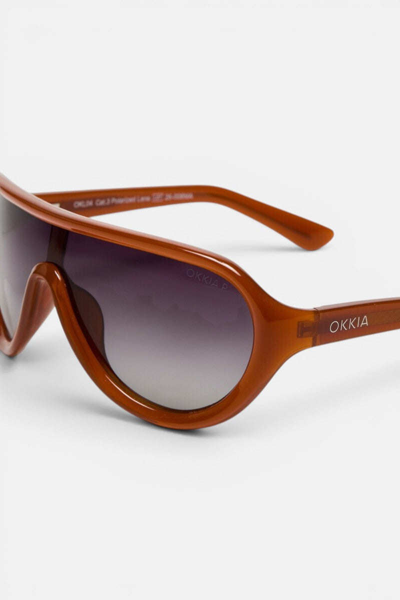Eine braune Kunststoffsonnenbrille mit großem, abgerundetem Rahmen und dunklen Verlaufsgläsern liegt vor einem schlichten weißen Hintergrund. Der Markenname „Okkia“ ist in weißer Schrift auf dem Bügel aufgedruckt, und auf der glatten Oberfläche der polarisierten Gläser sind Reflexionen zu sehen. Diese stilvolle Brille eignet sich mühelos als schickes Modeaccessoire für jeden modernen Look.