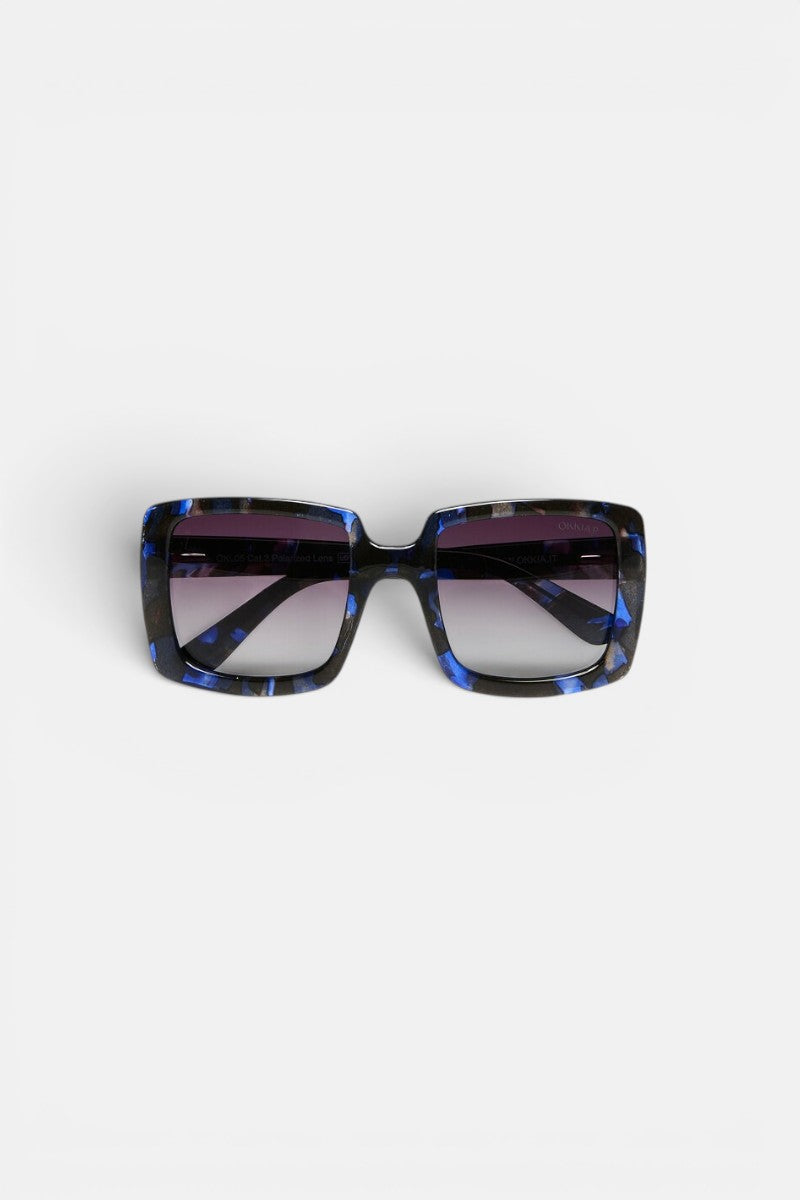 Die quadratische Sonnenbrille „ISCHIA“ von Okkia verfügt über eine schwarz-blau marmorierte Oversize-Fassung und dunkle, polarisierte Gläser mit Farbverlauf. Die Sonnenbrille wird mit der Vorderseite nach oben auf einem schlichten weißen Hintergrund präsentiert, wobei beide Bügel sichtbar sind und leicht nach außen gebogen sind.
