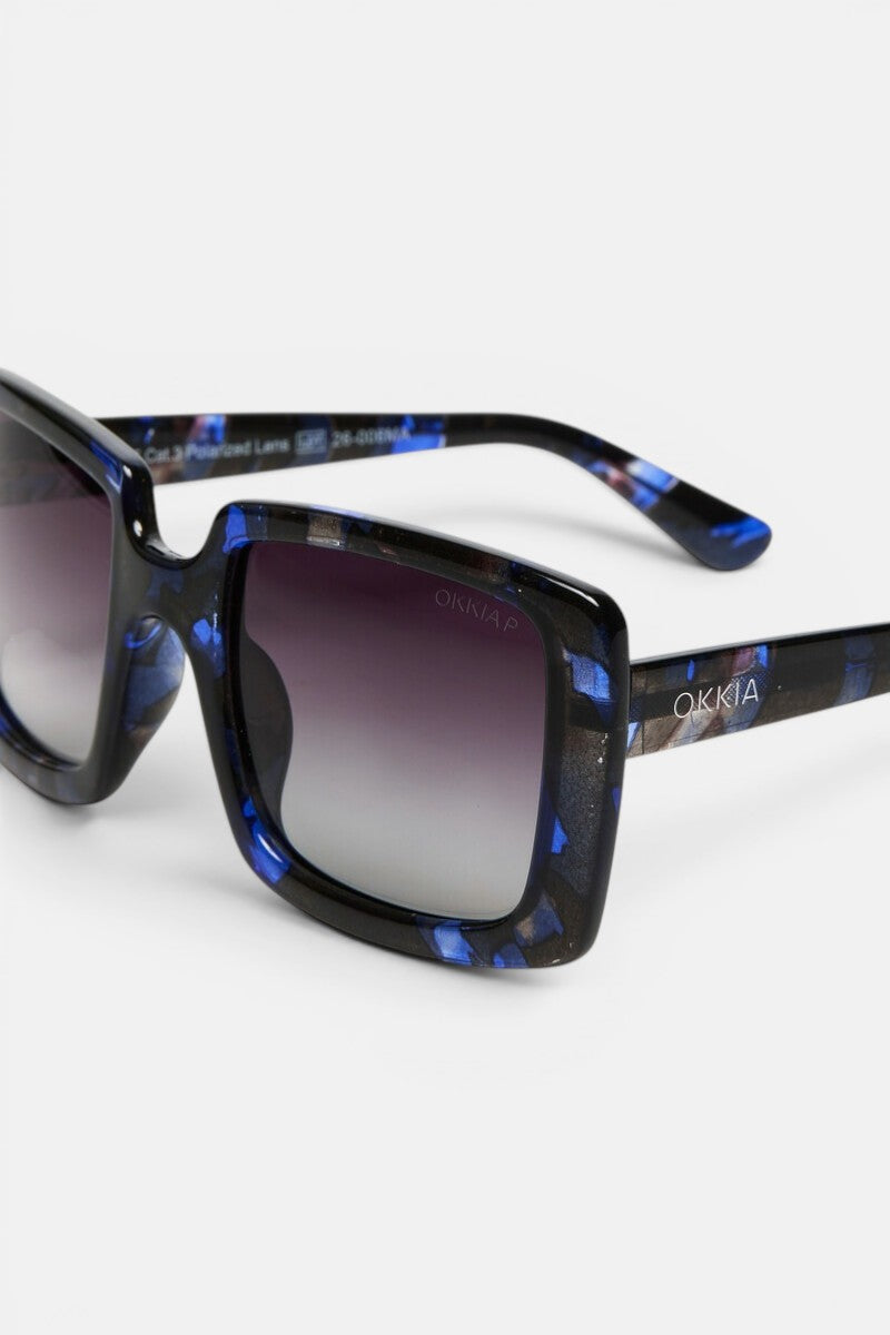 Eine Nahaufnahme der ISCHIA-Sonnenbrille zeigt das übergroße, eckige Brillengestell mit einem glänzenden, schwarz-blau marmorierten Rahmen, grauen, polarisierten Verlaufsglasen und dem in Weiß auf den Bügel aufgedruckten Markennamen „Okkia“. Das Bild präsentiert diese stilvolle Brille sowohl von der Seite als auch von vorne vor einem weißen Hintergrund.
