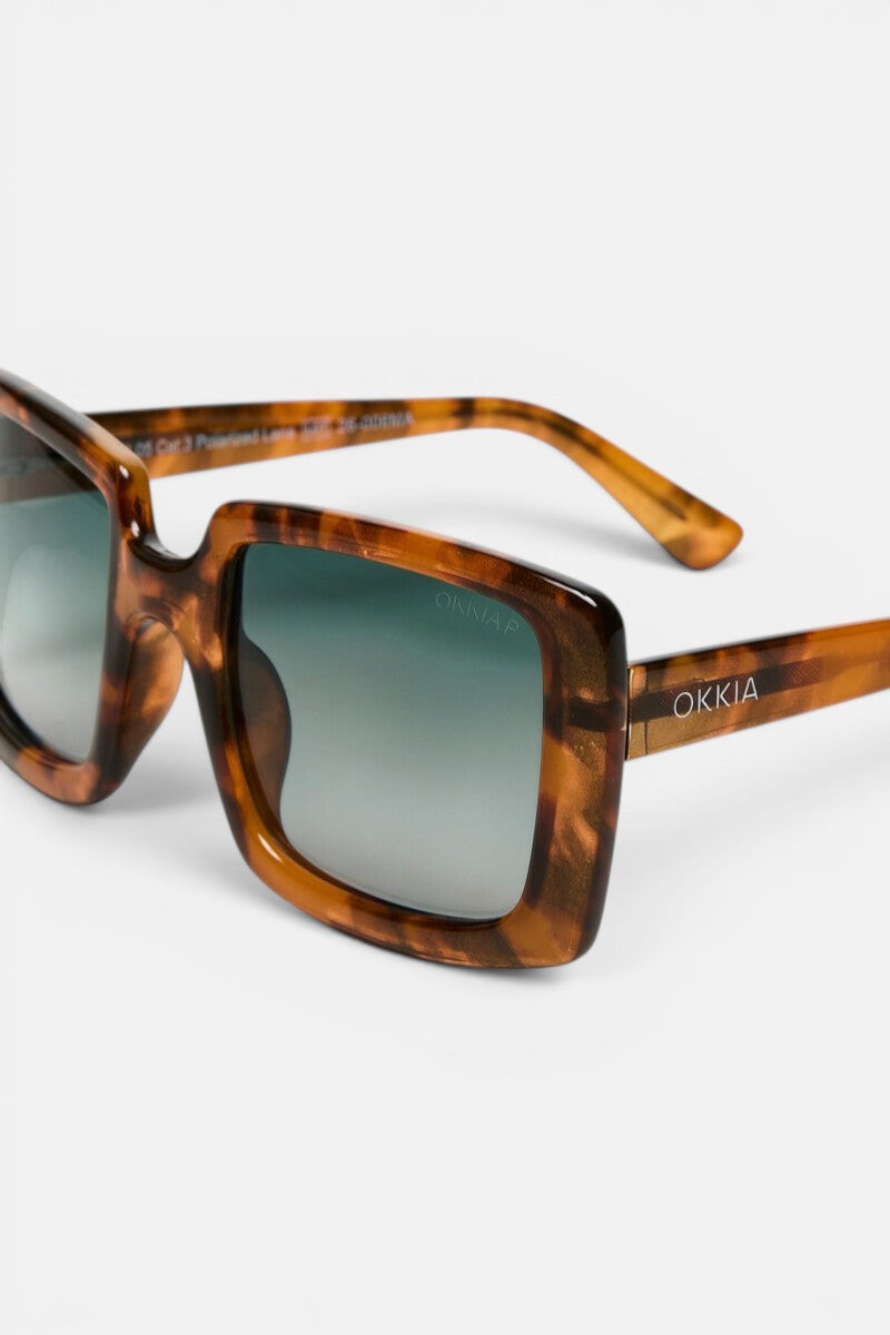 Nahaufnahme einer quadratischen Sonnenbrille im Schildpatt-Look mit dunklen, verlaufenden, polarisierten Gläsern. Die Sonnenbrille verfügt über einen breiten, braun-bernsteinfarben marmorierten Rahmen im Oversize-Design; auf dem linken Bügel ist „Okkia“ in Weiß aufgedruckt. Der Hintergrund ist schlicht weiß, wodurch diese stilvolle Okkia Club Sonnenbrille besonders gut zur Geltung kommt.
